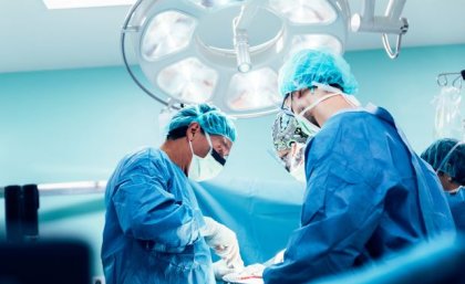 Image: Adobe. three people in blue gowns, surgical masks and gloves stand under a bright light looking down